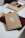 A journal with a simple design of a white branch and leaves on a brown cover rests on a table. Next to it, a box of stationery supplies, including tape and colored circles, is visible. A pen is tucked into the journal, which sits on top of a closed burgundy folder.
