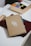 A journal with a simple design of a white branch and leaves on a brown cover rests on a table. Next to it, a box of stationery supplies, including tape and colored circles, is visible. A pen is tucked into the journal, which sits on top of a closed burgundy folder.