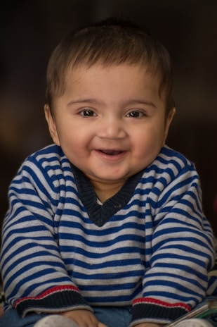 A smiling baby dressed in a striped sweater, captured against a soft, dark background. The baby's joyful expression is emphasized by its chubby cheeks and sparkling eyes.