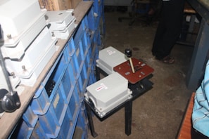 A cluttered storage area with rows of dusty blue storage bins stacked on metal shelves. In the foreground, there is a heavy-duty control panel with two white electrical boxes and a lever mechanism. The floor is made of bare concrete, and there is a person wearing dark pants standing near the setup.