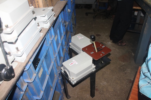 A cluttered storage area with rows of dusty blue storage bins stacked on metal shelves. In the foreground, there is a heavy-duty control panel with two white electrical boxes and a lever mechanism. The floor is made of bare concrete, and there is a person wearing dark pants standing near the setup.