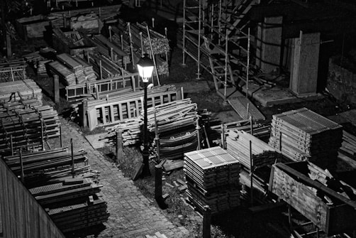 A black and white image depicts a construction site at night, illuminated by a single streetlamp. There are stacks of metal scaffolding and wooden planks neatly organized on the ground, with shadows cast by the light source adding depth and contrast. The area appears orderly yet deserted.