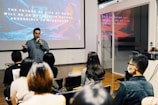 A speaker is presenting in front of an audience in a modern conference room. Behind the speaker, a large screen displays a slide with motivational text about the future. The audience is seated, some wearing masks, attentively listening to the presentation.