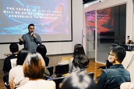 A speaker is presenting in front of an audience in a modern conference room. Behind the speaker, a large screen displays a slide with motivational text about the future. The audience is seated, some wearing masks, attentively listening to the presentation.