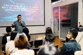 A speaker is presenting in front of an audience in a modern conference room. Behind the speaker, a large screen displays a slide with motivational text about the future. The audience is seated, some wearing masks, attentively listening to the presentation.