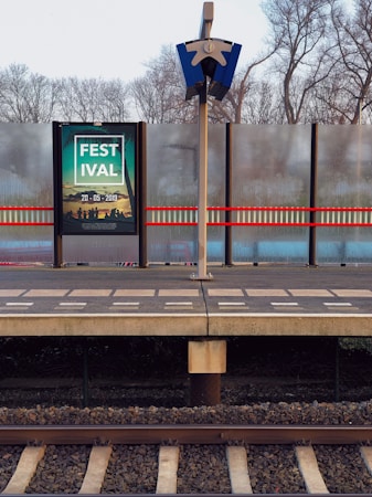 A train platform with a billboard displaying an advertisement for a festival dated 20-05-2019. The platform is bordered by a metal fence with frosted glass panels and there is a pole with a security or informational device attached to it. Surrounding trees are leafless, indicating late autumn or winter.