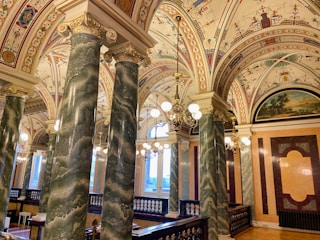 An ornately decorated interior featuring richly detailed ceilings with intricate designs and frescoes, supported by large green marble columns with gold accents. The room is illuminated by elegant chandeliers and has a warm, inviting atmosphere.