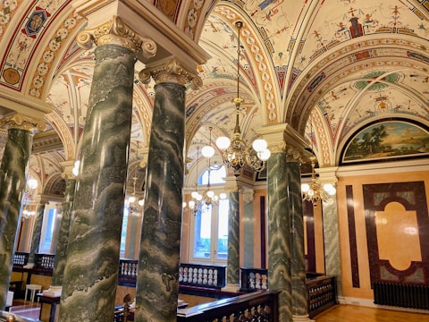 An ornately decorated interior featuring richly detailed ceilings with intricate designs and frescoes, supported by large green marble columns with gold accents. The room is illuminated by elegant chandeliers and has a warm, inviting atmosphere.