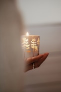 A hand gently holds a lit candle adorned with decorative tree designs. The candle emits a warm glow, highlighting intricate patterns on its surface. Light and shadows play softly around the scene, creating a peaceful ambiance.