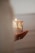 A hand gently holds a lit candle adorned with decorative tree designs. The candle emits a warm glow, highlighting intricate patterns on its surface. Light and shadows play softly around the scene, creating a peaceful ambiance.