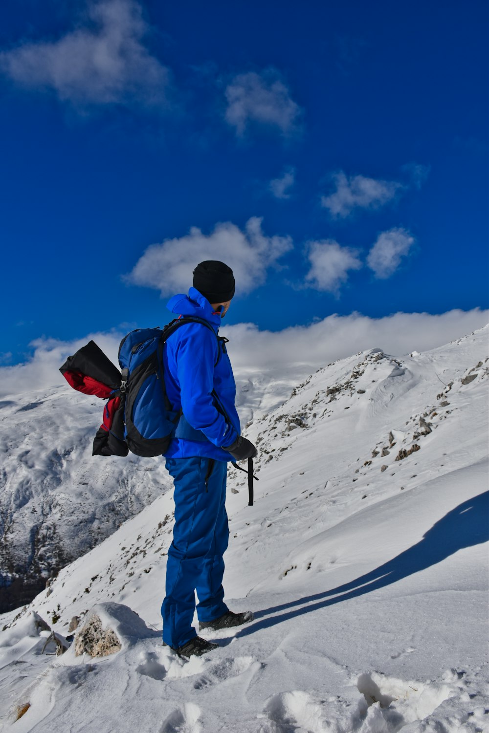 man in blue jacket and blue pants carrying black and red backpack standing on snow covered
