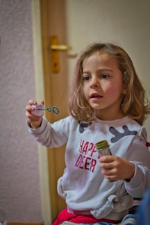 A young child with shoulder-length hair is indoors, holding a small wand for blowing bubbles in one hand and a small container in the other. The child is wearing a white sweatshirt with the words 'Happy Deer' in red and appears to be engaged and curious. The background features a closed wooden door and a textured wall.