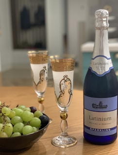 A bottle of sparkling wine labeled 'Latinium' stands next to two ornate, gold-rimmed champagne flutes. A bowl full of green and purple grapes sits nearby on a wooden table.