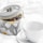 A decorative tin labeled 'TEA TIME' containing loose tea leaves is placed beside a plain white teacup and saucer on a soft white fabric background. The tin features a floral design with large blossoms in white and yellow.