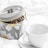 A decorative tin labeled 'TEA TIME' containing loose tea leaves is placed beside a plain white teacup and saucer on a soft white fabric background. The tin features a floral design with large blossoms in white and yellow.