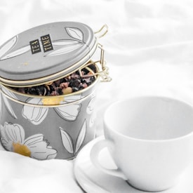 A decorative tin labeled 'TEA TIME' containing loose tea leaves is placed beside a plain white teacup and saucer on a soft white fabric background. The tin features a floral design with large blossoms in white and yellow.