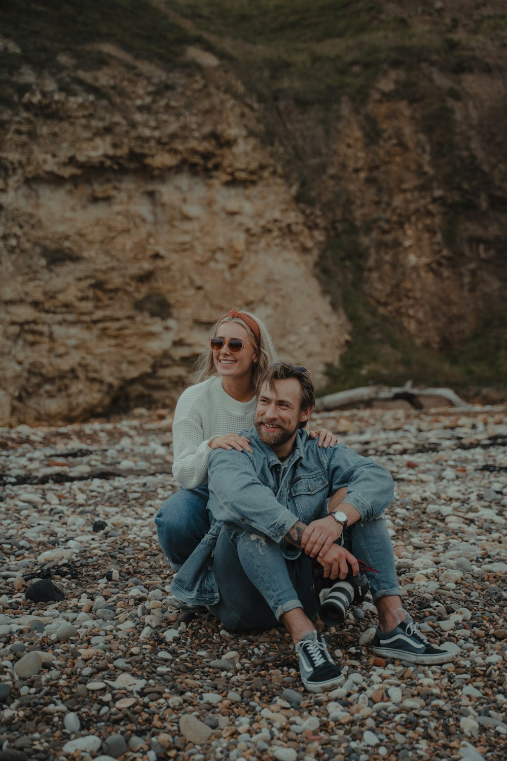 woman in white sweater sitting beside man in blue denim jeans on rocky ground during daytime