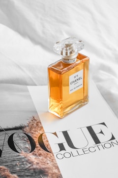 A Chanel No. 5 perfume bottle is placed on a magazine page that features the words 'VOGUE COLLECTION'. The background appears to be a white textured surface.