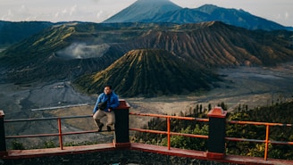 A person sitting on a railing with a panoramic view of volcanic mountains in the background, including a prominent crater. The landscape is lush and expansive, with varying shades of green and brown, under a clear sky.
