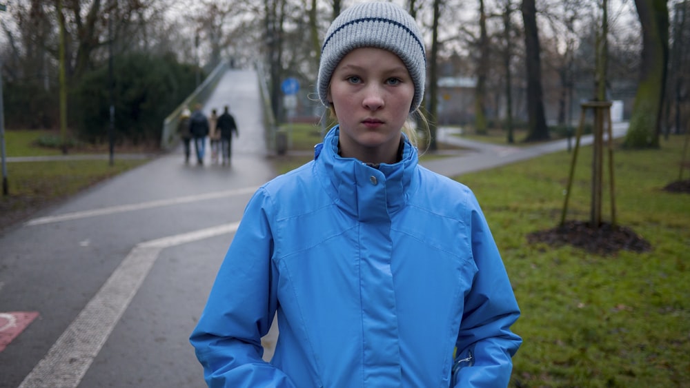 青いジャケットと灰色のニット帽をかぶった少年が昼間、歩道に立っている
