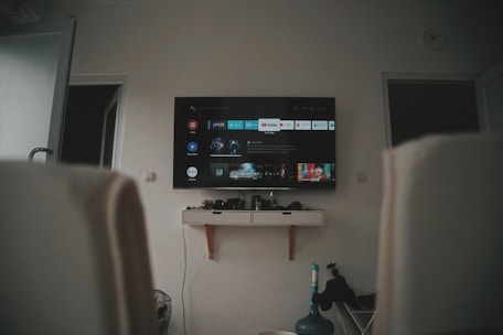 A living room setup featuring a wall-mounted TV displaying a streaming service interface. The room is dimly lit, with the TV as the main light source. Below the TV is a small shelf holding various objects. To the sides, there are partially visible chairs, and in the corner, there is a water bottle on the floor.