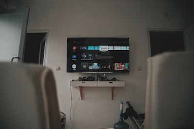 A living room setup featuring a wall-mounted TV displaying a streaming service interface. The room is dimly lit, with the TV as the main light source. Below the TV is a small shelf holding various objects. To the sides, there are partially visible chairs, and in the corner, there is a water bottle on the floor.