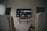 A living room setup featuring a wall-mounted TV displaying a streaming service interface. The room is dimly lit, with the TV as the main light source. Below the TV is a small shelf holding various objects. To the sides, there are partially visible chairs, and in the corner, there is a water bottle on the floor.