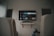 A living room setup featuring a wall-mounted TV displaying a streaming service interface. The room is dimly lit, with the TV as the main light source. Below the TV is a small shelf holding various objects. To the sides, there are partially visible chairs, and in the corner, there is a water bottle on the floor.