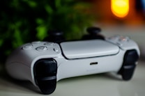 A white video game controller with black accents is lying on a white surface. The background features green leafy plants and a blurred warm light source, creating a cozy atmosphere.