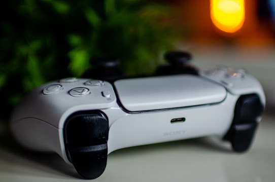A white video game controller with black accents is lying on a white surface. The background features green leafy plants and a blurred warm light source, creating a cozy atmosphere.