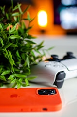 A gaming controller rests on a white surface beside a vibrant orange smartphone case and a leafy plant, with a warm, blurred light in the background.