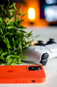 A gaming controller rests on a white surface beside a vibrant orange smartphone case and a leafy plant, with a warm, blurred light in the background.