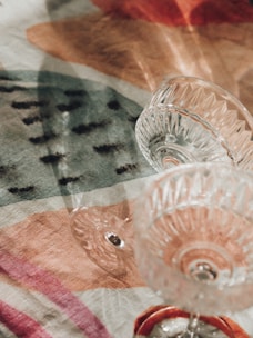 Glass cups with intricate designs are placed on a colorful, patterned fabric. The fabric features various shapes and colors, creating a visually appealing backdrop. The light reflects off the glass, casting subtle shadows on the fabric.