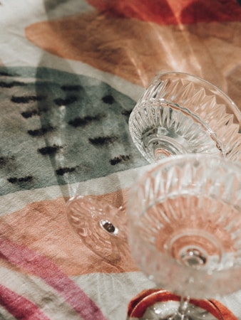 Glass cups with intricate designs are placed on a colorful, patterned fabric. The fabric features various shapes and colors, creating a visually appealing backdrop. The light reflects off the glass, casting subtle shadows on the fabric.
