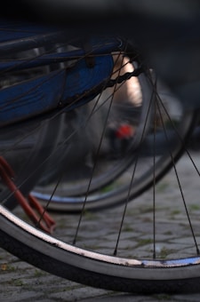 A close-up view of a bicycle wheel with the spokes and tire prominently visible. The background is blurred, suggesting other bicycles parked nearby. The scene is set on a cobblestone surface, and there is a metallic blue part of the bicycle frame in view, creating a play of light and shadow.