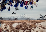 A breathtaking landscape featuring a distant snow-capped mountain surrounded by arid terrain, with several birds in mid-flight adding dynamism to the scene. In the foreground, colorful trinkets and evil eye ornaments hang from a tree branch, introducing a cultural element to the natural setting.