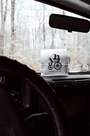 A paper bag with a logo featuring a bicycle and text 'Journey in a Cup' is placed on the dashboard of a vehicle. The view outside the windshield shows a forest with bare trees, indicating a winter or early spring setting. The interior of the vehicle includes a steering wheel and part of the central console.
