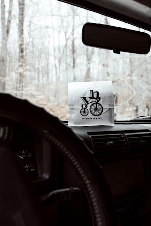 A paper bag with a logo featuring a bicycle and text 'Journey in a Cup' is placed on the dashboard of a vehicle. The view outside the windshield shows a forest with bare trees, indicating a winter or early spring setting. The interior of the vehicle includes a steering wheel and part of the central console.