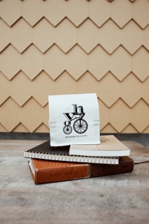 A small white paper bag with a black design of a bicycle and the words 'Journey in a Cup' sits atop two spiral notebooks and a thin book with a brown cover labeled 'Holy Bible'. The background features a beige shingle pattern, creating a textured backdrop.