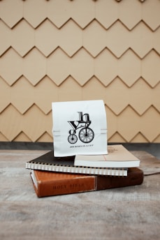A small white paper bag with a black design of a bicycle and the words 'Journey in a Cup' sits atop two spiral notebooks and a thin book with a brown cover labeled 'Holy Bible'. The background features a beige shingle pattern, creating a textured backdrop.