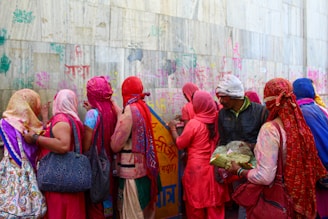 A group of people dressed in brightly colored traditional clothing are standing close together, writing or drawing on a wall covered in colorful chalk markings. The scene appears busy and vibrant, with most individuals facing away from the camera, revealing vividly decorated fabrics and headdresses.