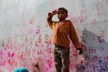 A child with a painted face stands against a colorful wall marked with handprints and various symbols. The child is wearing a brown jacket and appears to be posing with one hand raised to the forehead. The wall is covered in vibrant pink, red, and orange paint.