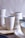 A collection of handcrafted ceramic cups and bowls, primarily in white and light blue hues, arranged on a wooden surface. The ceramics have a smooth texture with minimalistic and elegant designs.