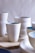 A collection of handcrafted ceramic cups and bowls, primarily in white and light blue hues, arranged on a wooden surface. The ceramics have a smooth texture with minimalistic and elegant designs.