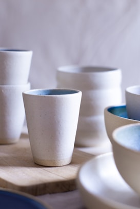 A collection of handcrafted ceramic cups and bowls, primarily in white and light blue hues, arranged on a wooden surface. The ceramics have a smooth texture with minimalistic and elegant designs.