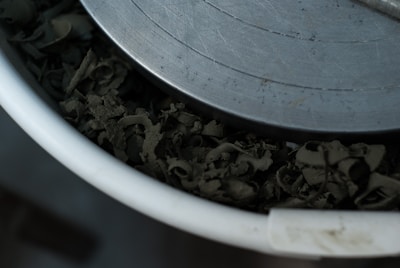A close-up view of a metal object placed over a collection of dark, curled shavings or curls inside a round white container. The metal surface shows circular grooves and a slightly worn appearance.