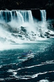 A large waterfall cascades down into a turbulent river below, with mist rising from the water's impact. A tour boat filled with people in blue raincoats approaches the falls, navigating through the churning water.