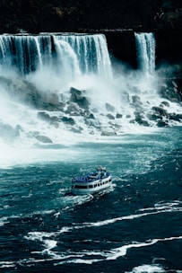 A large waterfall cascades down into a turbulent river below, with mist rising from the water's impact. A tour boat filled with people in blue raincoats approaches the falls, navigating through the churning water.
