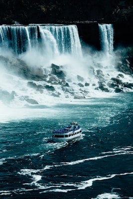 A large waterfall cascades down into a turbulent river below, with mist rising from the water's impact. A tour boat filled with people in blue raincoats approaches the falls, navigating through the churning water.