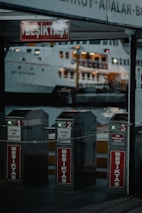 Three metallic turnstiles marked with 'Beşiktaş' are positioned at a ferry terminal entrance. Behind them, a large ferry boat is illuminated, docked at the harbor. The scene is set during the evening, with dim lighting and a slightly industrial ambiance.